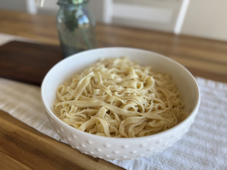 a white ceramic bowl filled with homemade pasta noodles on a wooden table with a neutral stripe table runner.