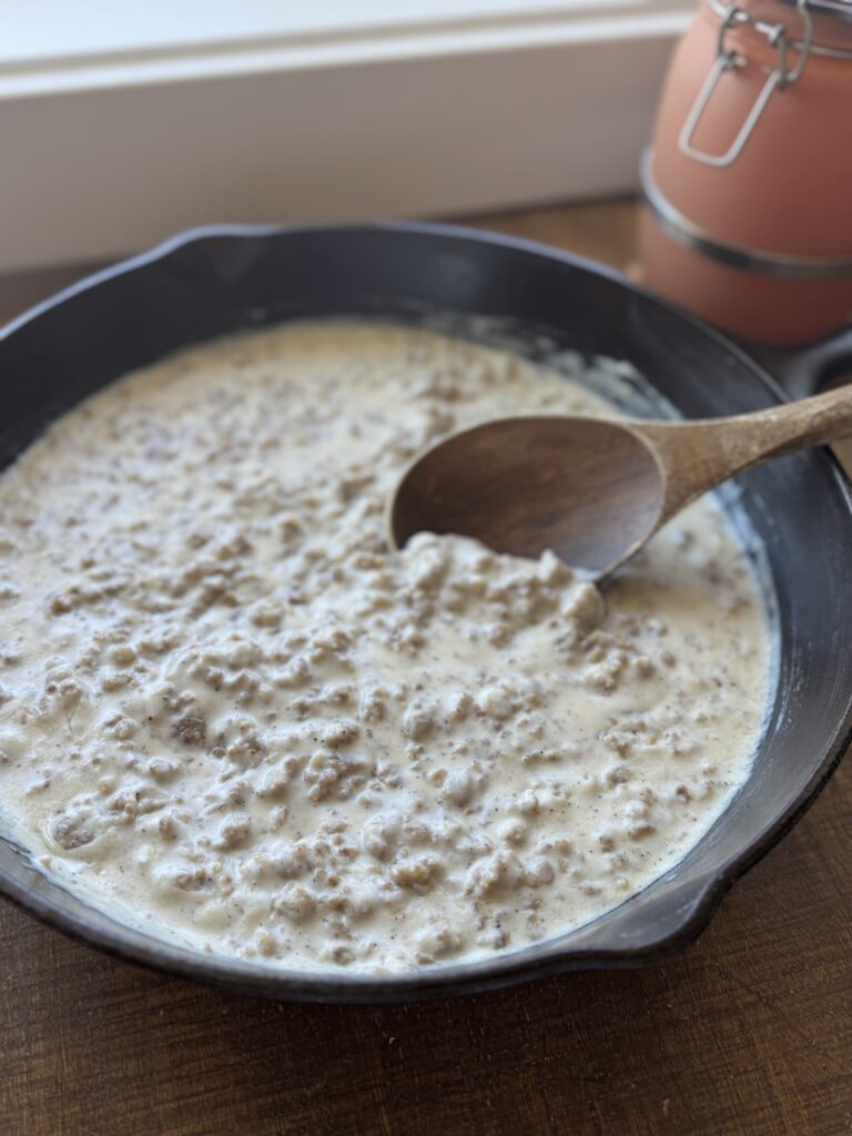 a Cast iron pan full of sausage gravy and a wooden spoon on a wooden table