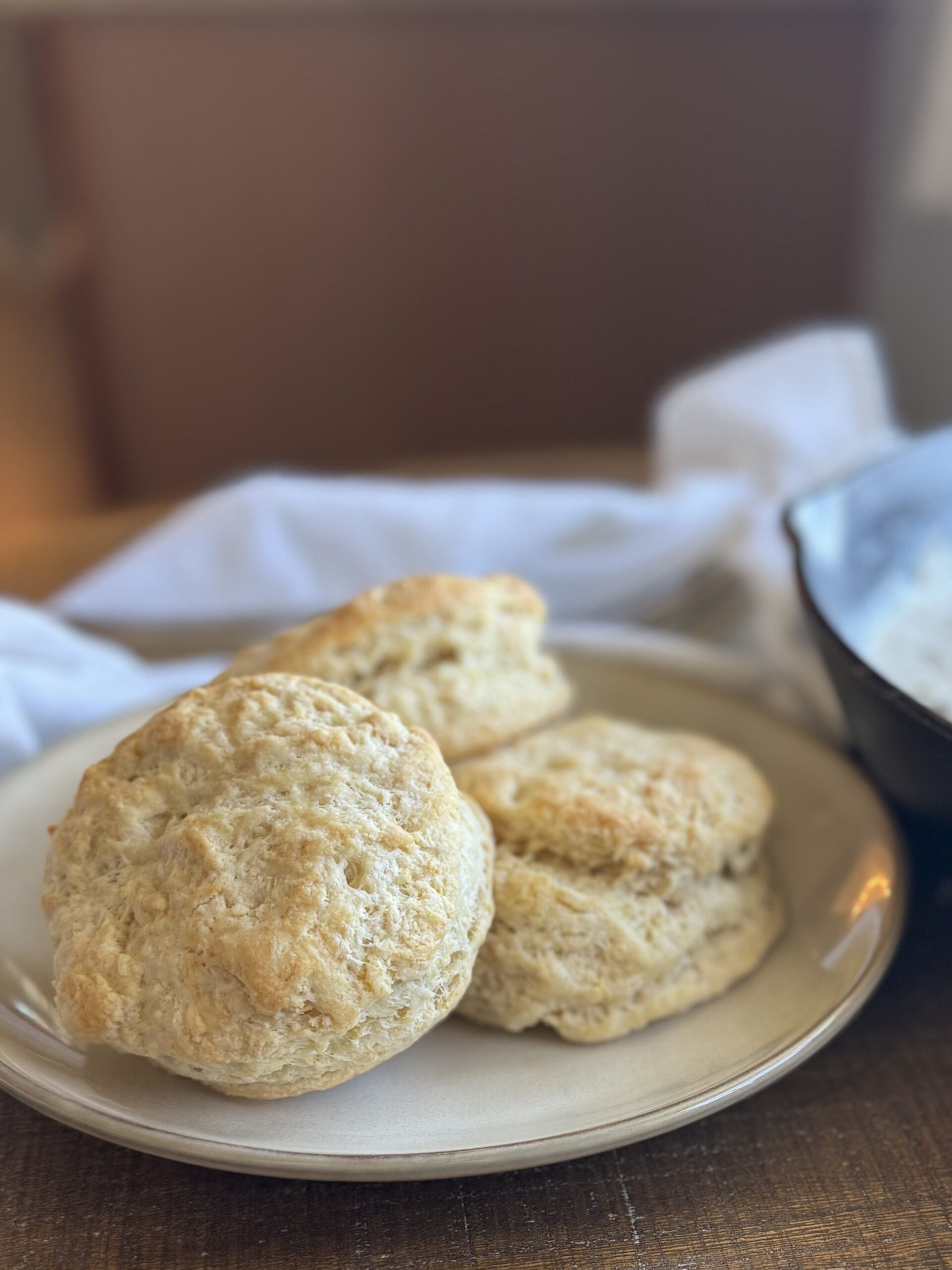 3 flaky biscuits on a plate with a cast iron pan near by and a white towel.