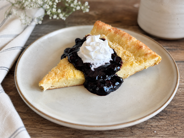 a slice of a dutch baby with berry sauce and whipped cream on a tan plate, on a wooden table with baby's breath and a linen towel next to it