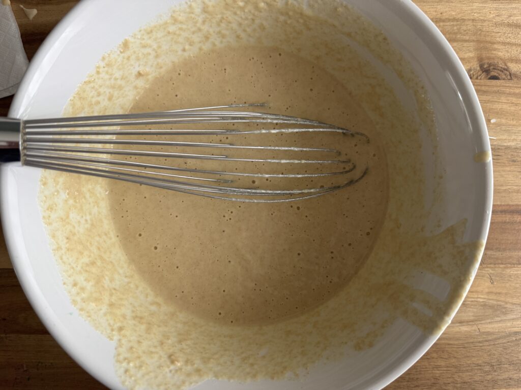 a white ceramic bowl on a wooden table with sourdough discard waffle batter and a whisk