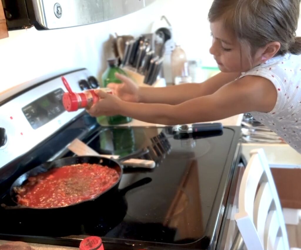 spices being added by a girl to spagetti sauce in a cast iron pan on a stove