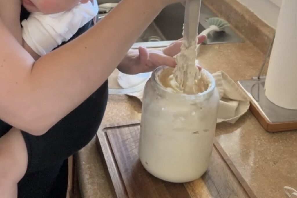 a mom stirring her sourdough starter with a wooden stirring stick on a counter.  