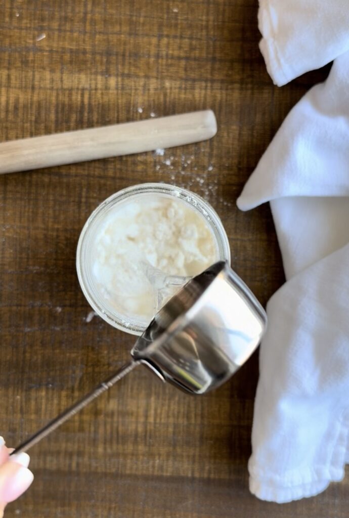 a sourdough starter being fed, with the water being added in with a measuring cup