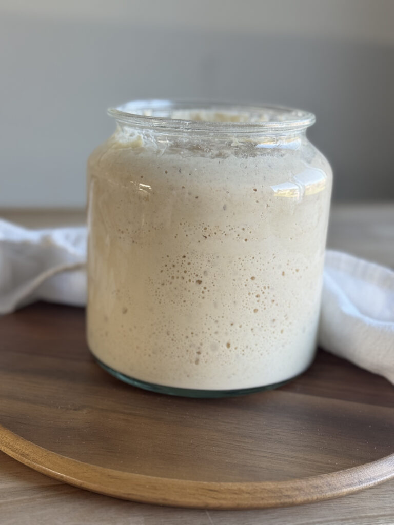a jar of sourdough starter on a wooden cutting board on a table with a white tea towel