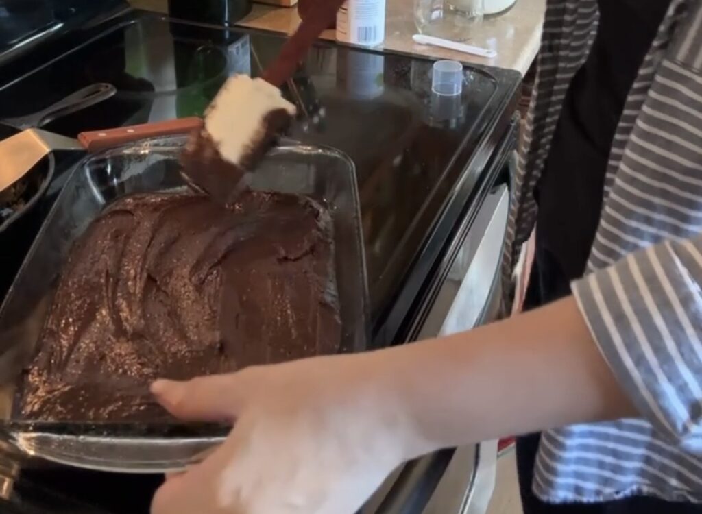 a woman spreading the brownie batter in the pan on top of a stove with a spatula