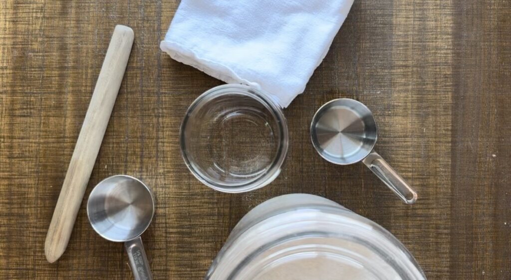 a overhead shot of supplies needed to make a sourdough starter, jar, water, stir stick, towel, flour